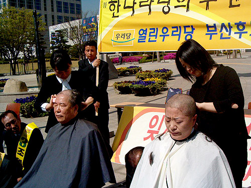 열린우리당 부산시선거위원회 조성래, 윤원호 공동위원장(사진왼쪽부터)이 삭발식을 행하고 있다. 삭발식이 진행되는 동안 총선후보들은 주위에 무릎을 꿇고 고개를 숙이고 있다. 